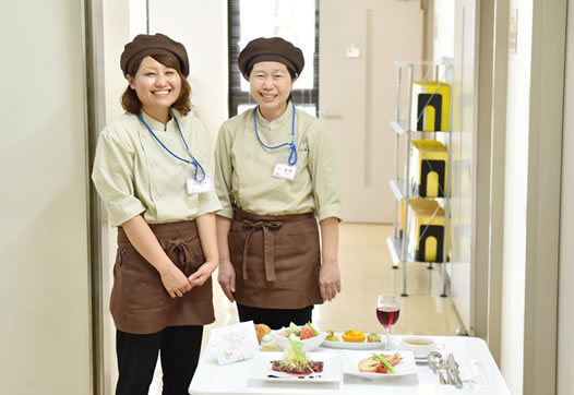 写真：テーブルに置かれた豪華な食事と、その後ろに立っている二人の女性スタッフ