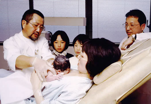 写真：生まれたばかりの赤ちゃんを母親の胸の上に置いている医者、その横には2人の女の子と父親